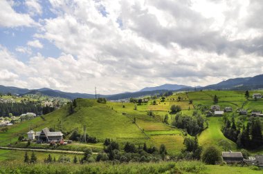 gökyüzünde parlak bulutlar ve güneş ışığı ile güzel bir dağ manzara, kabarık yeşil ladin, ve dağ yamaçlarında konut binaları ile tarifsiz güzellik glade. fantastik açık arka plan.