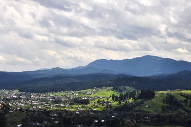 gökyüzünde parlak bulutlar ve güneş ışığı ile güzel bir dağ manzara, kabarık yeşil ladin, ve dağ yamaçlarında konut binaları ile tarifsiz güzellik glade. fantastik açık arka plan.