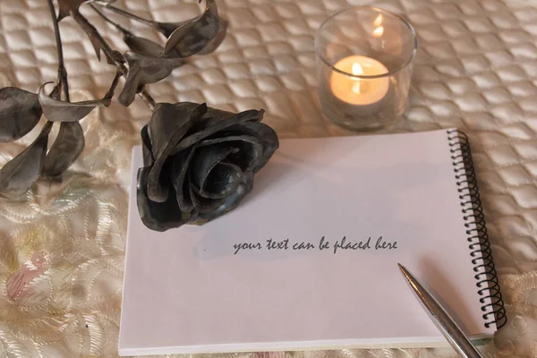 Hygge style. A lit candle illuminates a notebook on which lies a silver pen. a forged rose, as a symbol of eternal love, lies nearby. vintage style message template
