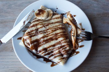 Kahvaltıda muz, ceviz ve çikolatalı krep. Çatal bıçak takımı ile seramik beyaz plaka. Servis tablosu