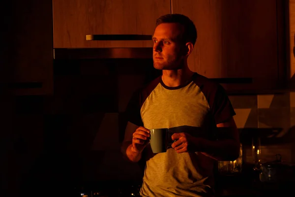 A man standing in front of a window in sunset lighting is drinking coffee in his kitchen