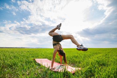 Yeşil çimenlerin üzerinde güzel bir kız model yoga yapıyor. Yeşil çimenlikteki güzel genç bir kadın akrobatik öğeler sergiliyor. Siyahlı esnek jimnastikçi ikiye bölünmüş amuda kalkar