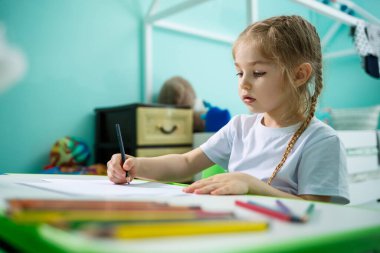 Beyaz tişörtlü bir kız masasında oturmuş renkli kalemlerle resim çiziyor.