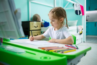 Beyaz tişörtlü bir kız masasında oturmuş renkli kalemlerle resim çiziyor.