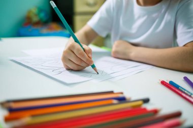 Küçük kız duvarın arka planında bir masada otururken resim çizer.