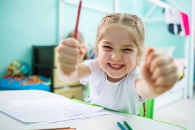 Küçük kız duvarın arka planında bir masada otururken resim çizer.
