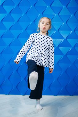 Yedi yaşında, ceketli, mavi arkaplanda bezelyeler olan sarışın bir kız dans ediyor, gülüyor, neşeli çocuklar, mutlu bir çocukluk geçiriyor.