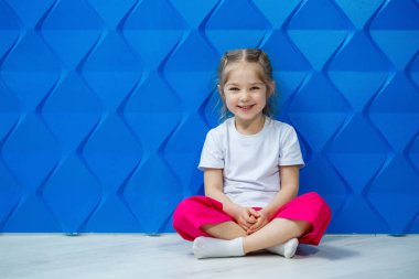 Mavi arka planda beyaz tişörtlü at kuyruklu küçük kız. Nilüfer pozisyonunda oturur ve gülümser ve ellerini sallar..