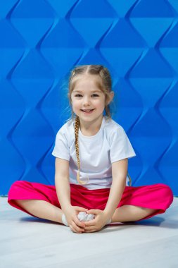 Mavi arka planda beyaz tişörtlü at kuyruklu küçük kız. Nilüfer pozisyonunda oturur ve gülümser ve ellerini sallar..