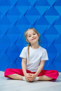 Mavi arka planda beyaz tişörtlü at kuyruklu küçük kız. Nilüfer pozisyonunda oturur ve gülümser ve ellerini sallar..