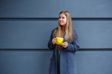 Gri montlu güzel sarışın kadın soğuk bir sonbahar gününde sarı fincandan sıcak çay içer. Kafedeki terasta nefis sıcak bir içecek.