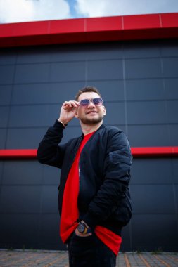 Gözlüklü, kırmızı tişörtlü, ceketli, sakallı genç bir adam gri bir binanın yanında poz veriyor. Çekici bir adam bir yaz günü şehirde dinleniyor. Erkek modası.