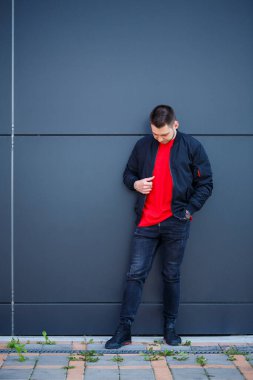 Sıradan giyinmiş genç bir adam, siyah ceketli, kot pantolonlu, kırmızı tişörtlü. Gri bir duvara karşı.
