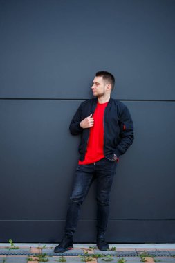 Sıradan giyinmiş genç bir adam, siyah ceketli, kot pantolonlu, kırmızı tişörtlü. Gri bir duvara karşı.