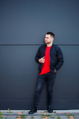 Sıradan giyinmiş genç bir adam, siyah ceketli, kot pantolonlu, kırmızı tişörtlü. Gri bir duvara karşı.