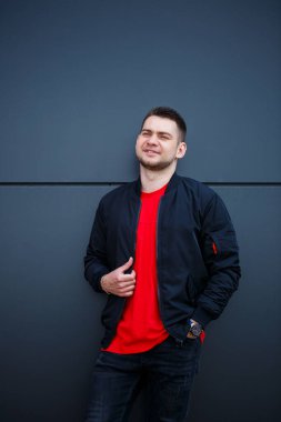 Sokak tarzı giyinmiş genç bir adam gri bir duvara karşı duruyor, siyah ceket ve kırmızı tişört giyiyor. Tarz sahibi adamın orkestrası senin promosyon mesajının arka planı..
