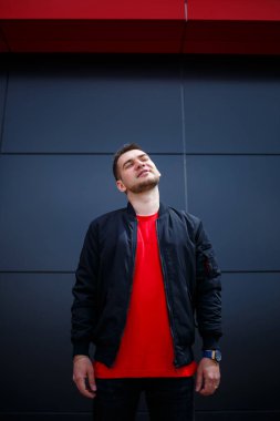 Sokak tarzı giyinmiş genç bir adam gri bir duvara karşı duruyor, siyah ceket ve kırmızı tişört giyiyor. Tarz sahibi adamın orkestrası senin promosyon mesajının arka planı..