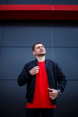 Sokak tarzı giyinmiş genç bir adam gri bir duvara karşı duruyor, siyah ceket ve kırmızı tişört giyiyor. Tarz sahibi adamın orkestrası senin promosyon mesajının arka planı..