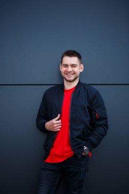 Sokak tarzı giyinmiş genç bir adam gri bir duvara karşı duruyor, siyah ceket ve kırmızı tişört giyiyor. Tarz sahibi adamın orkestrası senin promosyon mesajının arka planı..