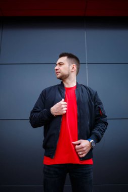 Sokak tarzı giyinmiş genç bir adam gri bir duvara karşı duruyor, siyah ceket ve kırmızı tişört giyiyor. Tarz sahibi adamın orkestrası senin promosyon mesajının arka planı..