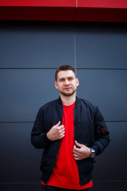 Sokak tarzı giyinmiş genç bir adam gri bir duvara karşı duruyor, siyah ceket ve kırmızı tişört giyiyor. Tarz sahibi adamın orkestrası senin promosyon mesajının arka planı..