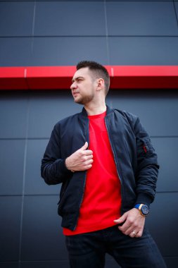 Sokak tarzı giyinmiş genç bir adam gri bir duvara karşı duruyor, siyah ceket ve kırmızı tişört giyiyor. Tarz sahibi adamın orkestrası senin promosyon mesajının arka planı..