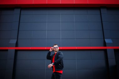 Günlük giysili, kırmızı tişörtlü, siyah ceketli gri duvarlı bir sokakta gülümseyen genç bir Avrupalı. Modern bir adam.