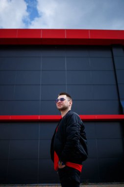 Gözlüklü, kırmızı tişörtlü, ceketli, sakallı genç bir adam gri bir binanın yanında poz veriyor. Çekici bir adam bir yaz günü şehirde dinleniyor. Erkek modası.