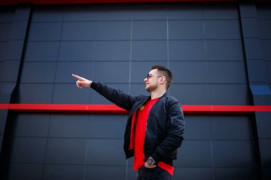 Gözlüklü, kırmızı tişörtlü, ceketli, sakallı genç bir adam gri bir binanın yanında poz veriyor. Çekici bir adam bir yaz günü şehirde dinleniyor. Erkek modası.