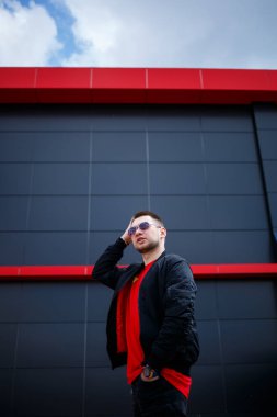Gözlüklü, kırmızı tişörtlü, ceketli, sakallı genç bir adam gri bir binanın yanında poz veriyor. Çekici bir adam bir yaz günü şehirde dinleniyor. Erkek modası.
