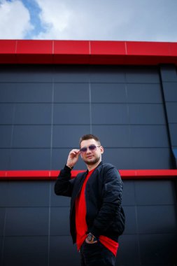 Gözlüklü, kırmızı tişörtlü, ceketli, sakallı genç bir adam gri bir binanın yanında poz veriyor. Çekici bir adam bir yaz günü şehirde dinleniyor. Erkek modası.