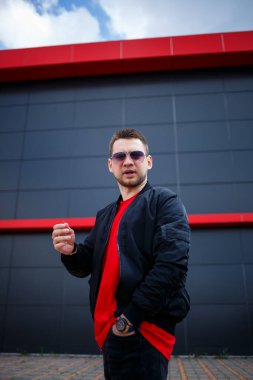 Gözlüklü, kırmızı tişörtlü, ceketli, sakallı genç bir adam gri bir binanın yanında poz veriyor. Çekici bir adam bir yaz günü şehirde dinleniyor. Erkek modası.