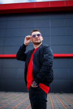 Gözlüklü, kırmızı tişörtlü, ceketli, sakallı genç bir adam gri bir binanın yanında poz veriyor. Çekici bir adam bir yaz günü şehirde dinleniyor. Erkek modası.