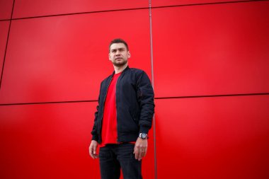Havalı, güneş gözlüklü, şık kırmızı tişörtlü, siyah ceketli genç bir adam şehirde kırmızı bir binanın yanında duruyor. Çekici, modern adam. Şık yaz erkek kıyafetleri. Sokak tarzı