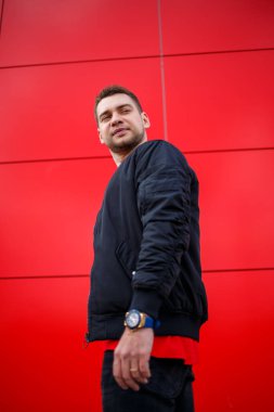 Havalı, güneş gözlüklü, şık kırmızı tişörtlü, siyah ceketli genç bir adam şehirde kırmızı bir binanın yanında duruyor. Çekici, modern adam. Şık yaz erkek kıyafetleri. Sokak tarzı