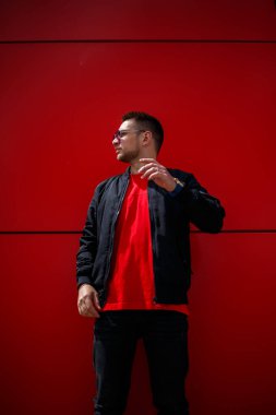 Havalı, güneş gözlüklü, şık kırmızı tişörtlü, siyah ceketli genç bir adam şehirde kırmızı bir binanın yanında duruyor. Çekici, modern adam. Şık yaz erkek kıyafetleri. Sokak tarzı