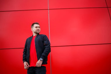 Havalı, güneş gözlüklü, şık kırmızı tişörtlü, siyah ceketli genç bir adam şehirde kırmızı bir binanın yanında duruyor. Çekici, modern adam. Şık yaz erkek kıyafetleri. Sokak tarzı