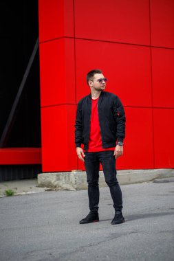 Sakallı, neşeli bir genç adam. Tişörtünde olumlu bir gülümseme var. Siyah bir ceketle, şehirde Red Metal bir binanın yanında poz veriyor. Sokaktaki mutlu şehirli adam. Erkek kıyafetleri.