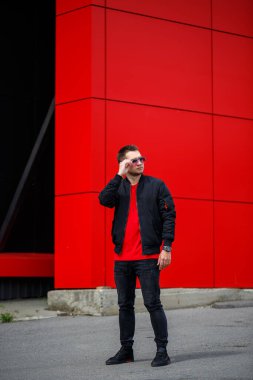 Sakallı, neşeli bir genç adam. Tişörtünde olumlu bir gülümseme var. Siyah bir ceketle, şehirde Red Metal bir binanın yanında poz veriyor. Sokaktaki mutlu şehirli adam. Erkek kıyafetleri.