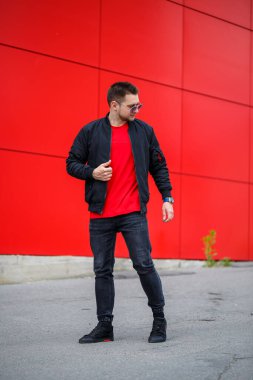 Kırmızı binanın yakınında güneşli bir günde güneş gözlüklü, klasik kot pantolonlu şık bir genç adam poz veriyor. Sokaktaki modern adam. Gençlik tarzı