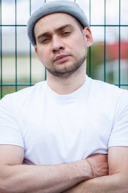Tarz sahibi genç bir adam, beyaz bir tişört ve gri bir şapka giymiş bir adam bir futbol sahasının arka planında duruyor. Şehir tarzı kıyafetler, modern moda imajı. Erkek modası