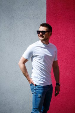 Beyaz tişört giymiş, gri ve kırmızı duvar arkasında duran yakışıklı bir adamın portresi. Şehir tarzı kıyafetler, modern moda imajı. Erkek modası