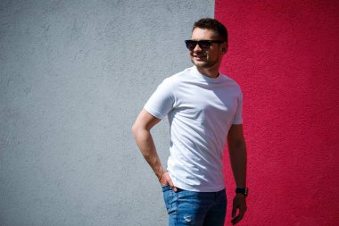 Beyaz tişört giymiş, gri ve kırmızı duvar arkasında duran yakışıklı bir adamın portresi. Şehir tarzı kıyafetler, modern moda imajı. Erkek modası