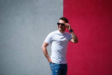Beyaz tişört giymiş, gri ve kırmızı duvar arkasında duran yakışıklı bir adamın portresi. Şehir tarzı kıyafetler, modern moda imajı. Erkek modası