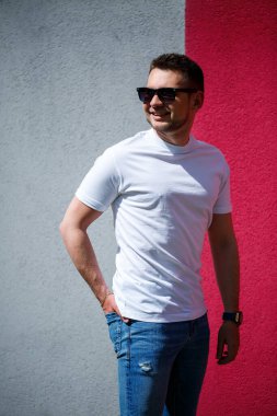 Beyaz tişört giymiş, gri ve kırmızı duvar arkasında duran yakışıklı bir adamın portresi. Şehir tarzı kıyafetler, modern moda imajı. Erkek modası