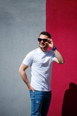Tarz sahibi genç bir adamın portresi, beyaz bir tişört giymiş gri ve kırmızı bir duvar arkasında duran bir adam. Şehir tarzı kıyafetler, modern moda imajı. Erkek modası