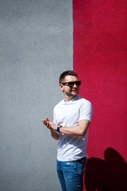 Tarz sahibi genç bir adam, beyaz bir tişört giymiş gri ve kırmızı bir duvar arkasında duran bir adam. Şehir tarzı kıyafetler, modern moda imajı. Erkek modası