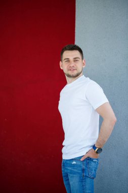 Tarz sahibi genç bir adam, beyaz bir tişört giymiş gri ve kırmızı bir duvar arkasında duran bir adam. Şehir tarzı kıyafetler, modern moda imajı. Erkek modası