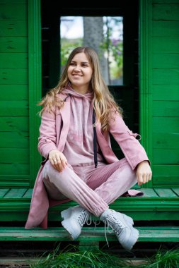 Avrupa görünümlü sarı saçlı şık genç kız merdivenlerde oturuyor, pembe bir takım elbise giyiyor, modern kadın modası, gündelik kıyafetler.