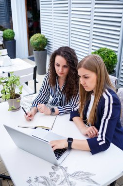 İki başarılı genç iş kadını yöneticisi bir dizüstü bilgisayar ve bir defterle bir masada oturmuş yeni bir geliştirme projesi üzerinde çalışıyor, bayan öğrenciler bilgisayarlarının çalışması üzerine bir rapor yazıyorlar.